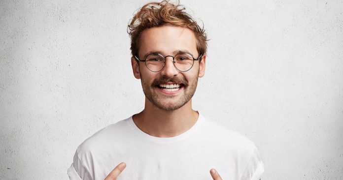Smiling young male indicates at blank t shirt, has happy expression, glad to advertize new clothes, isolated over concrete background. Unshaven cheerful hipster points at new outfit. Design, clothing