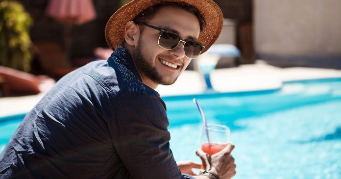 Mężczyzna w okularach przeciwsłonecznych i kapeluszu pije koktajl, siedząc w pobliżu basenu.