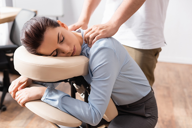 Masażysta masuje ramiona kobiety z zamkniętymi oczami, siedzącej na fotelu do masażu